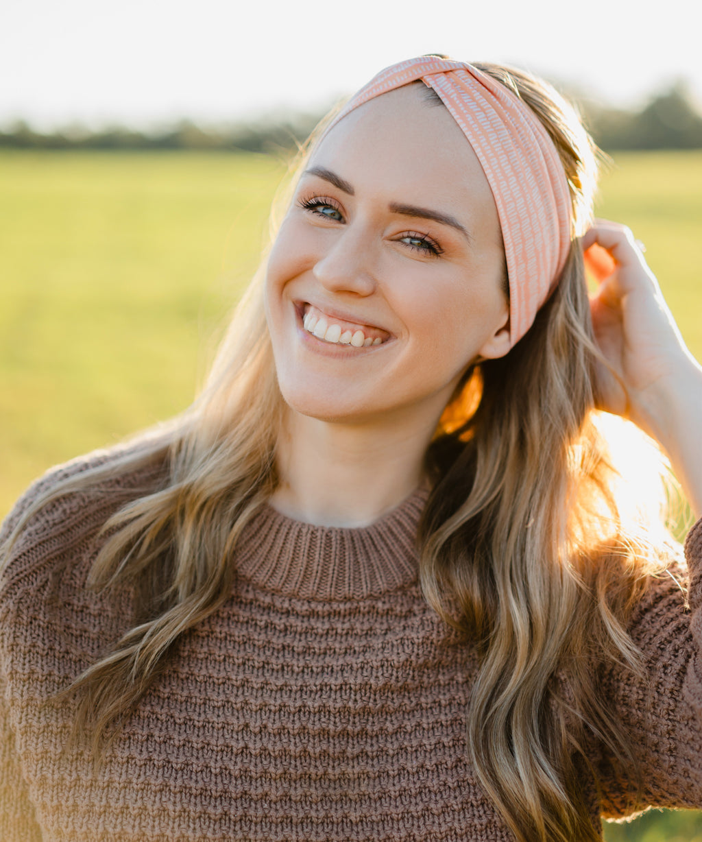 Woman smiling, wearing a Spiced Peach headband, showcasing its versatility and comfort, ideal for workouts or casual wear, made from soft, stretchy poly-knit blend.