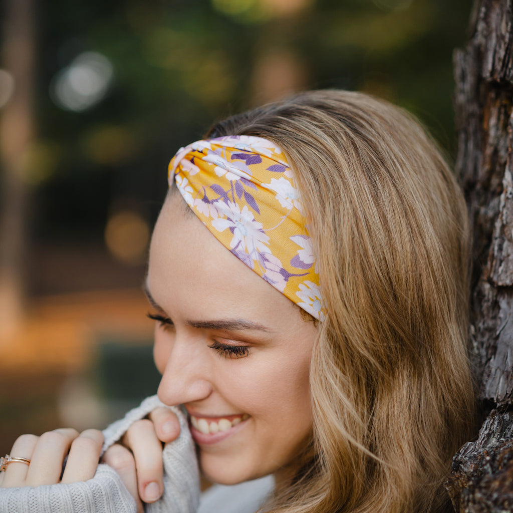 Autumn Daisy headband with purple and white flowers, worn by smiling woman, showcasing its versatile 3-in-1 twist style and cozy, stretchy poly-knit fabric.