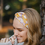 Autumn Daisy headband with purple and white flowers, worn by smiling woman, showcasing its versatile 3-in-1 twist style and cozy, stretchy poly-knit fabric.