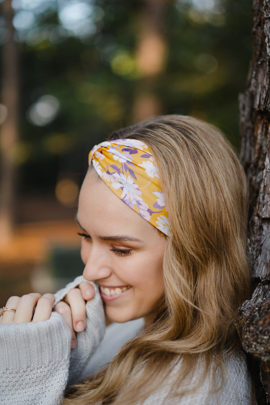 Autumn Daisy headband with purple and white flowers, worn by smiling woman, showcasing its versatile 3-in-1 twist style and cozy, stretchy poly-knit fabric.