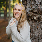 Autumn Daisy headband with purple and white flowers, worn by smiling woman, showcasing its versatile 3-in-1 twist style and cozy, stretchy poly-knit fabric.