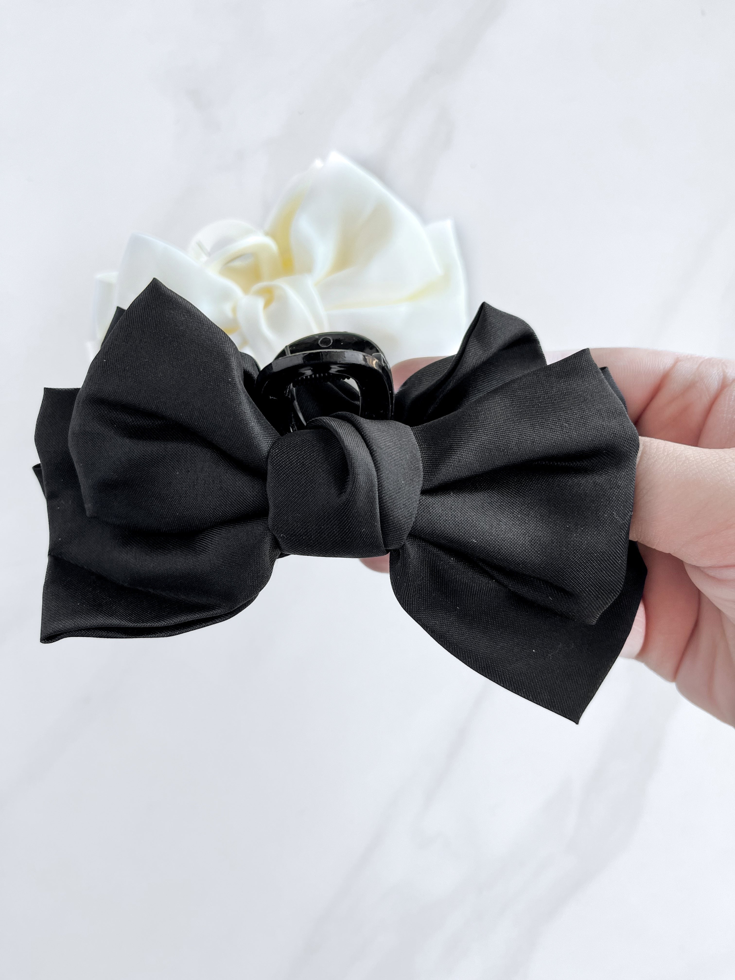 A hand holding a Classy Chic Lilac Clip, a perfect claw clip for all hair types and textures, made of silky black satin material with a hinge closure.
