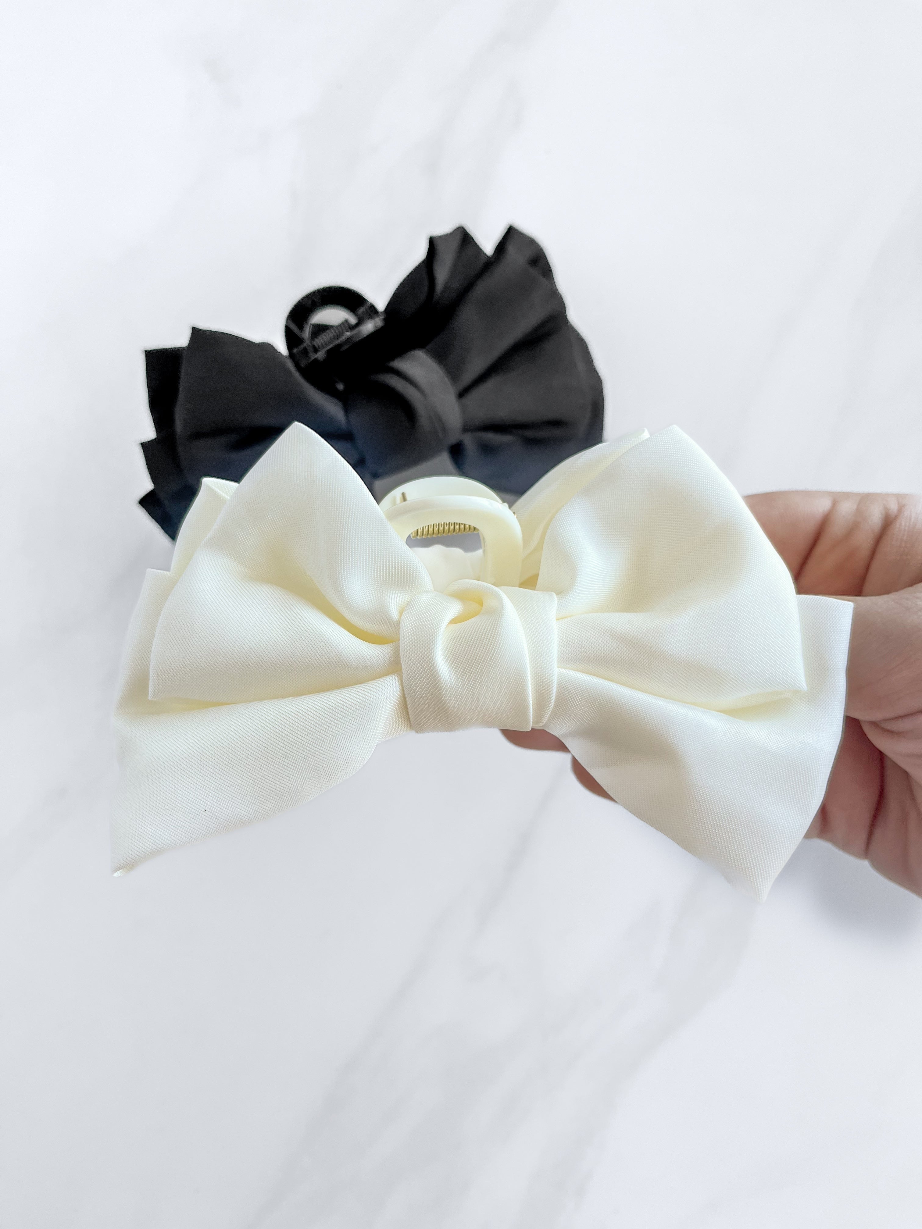 A hand holding a Classy Chic Lilac Clip, a perfect claw clip for all hair types and textures, made of silky white satin material with a hinge closure.