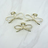 A group of shiny gold metal Invisible String Lilac Clips in a bow tie shape with a hinge closure, 3.5 inches long. Perfect for all hair types. Hair-out-of-face approved.