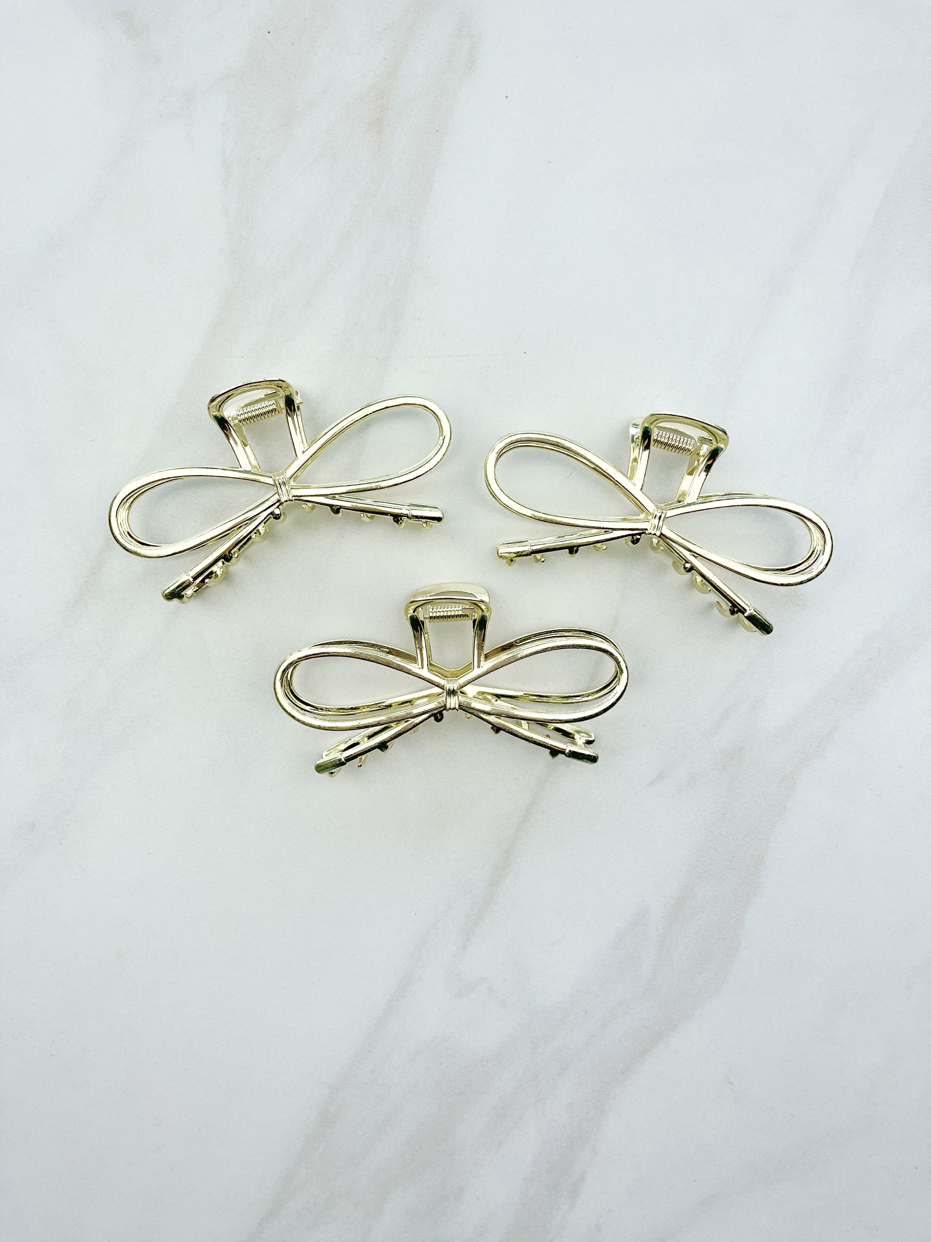 A group of shiny gold metal Invisible String Lilac Clips in a bow tie shape with a hinge closure, 3.5 inches long. Perfect for all hair types. Hair-out-of-face approved.