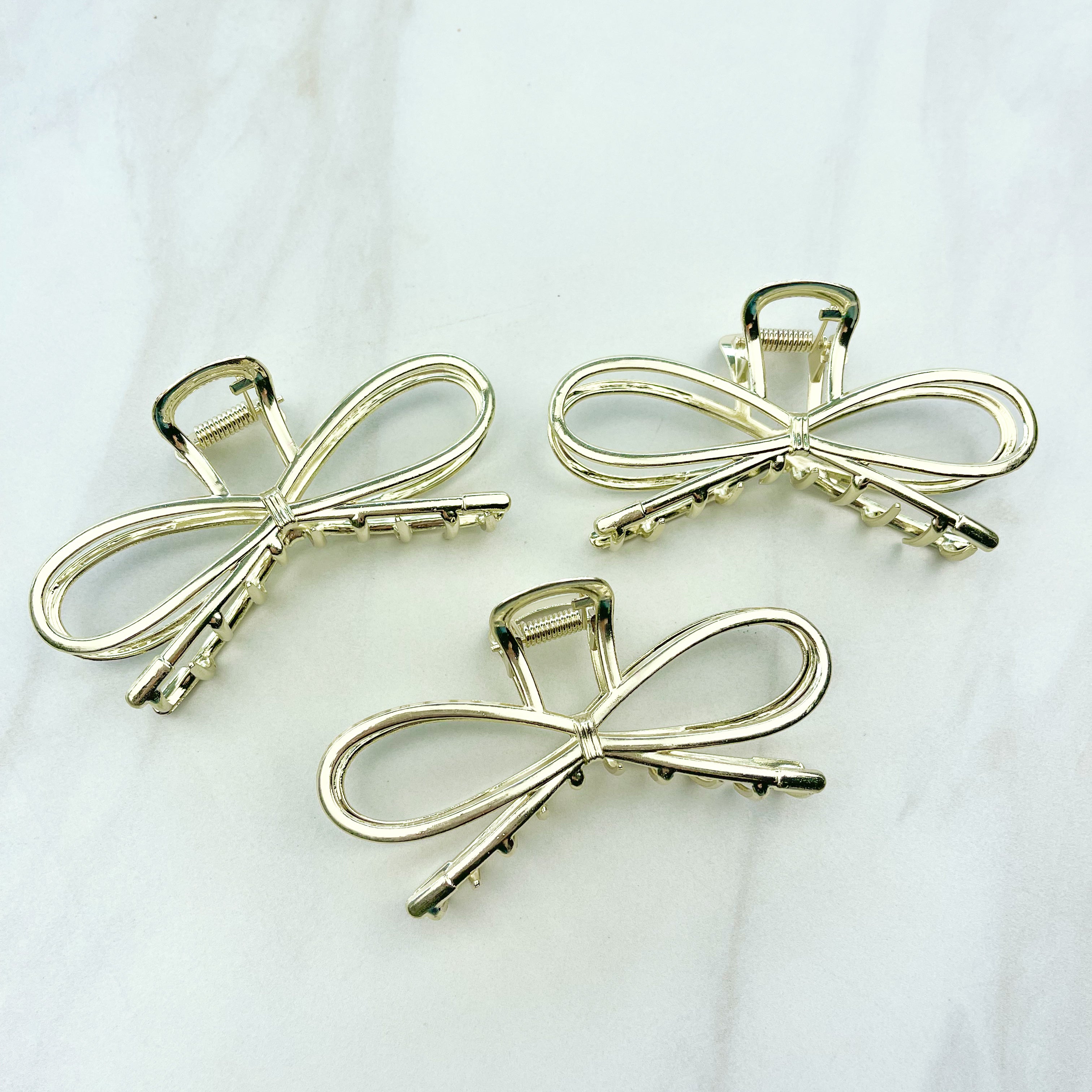 A group of shiny gold metal Invisible String Lilac Clips in a bow tie shape with a hinge closure, 3.5 inches long. Perfect for all hair types. Hair-out-of-face approved.