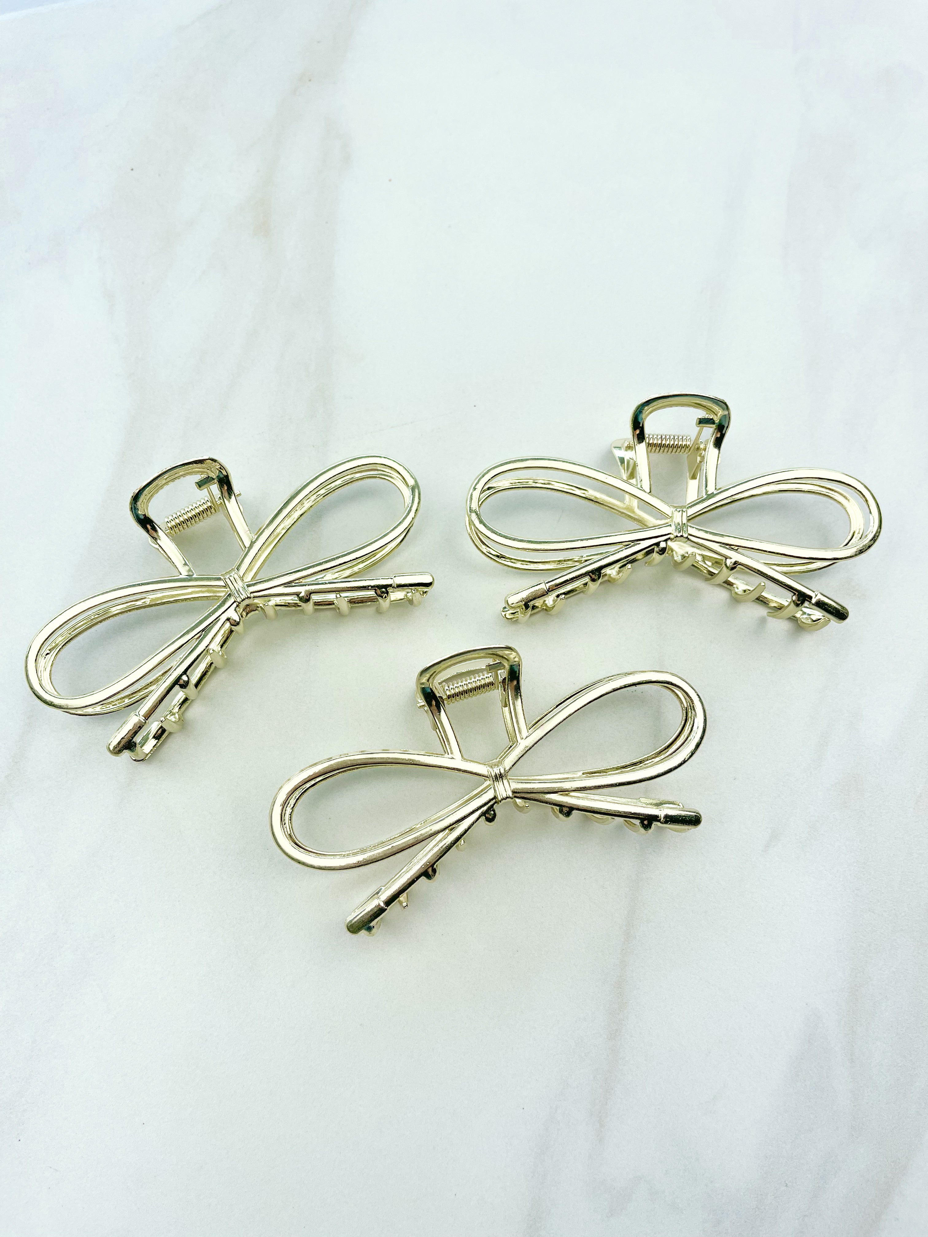 A group of shiny gold metal Invisible String Lilac Clips in a bow tie shape with a hinge closure, 3.5 inches long. Perfect for all hair types. Hair-out-of-face approved.
