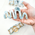 A group of zig zag-shaped blue marble hair clips with gold trim, held in a woman's hand to show the large size.