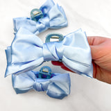 A hand holding a Classy Chic Lilac Clip, a perfect claw clip for all hair types and textures, made of blue silky satin material with a hinge closure.