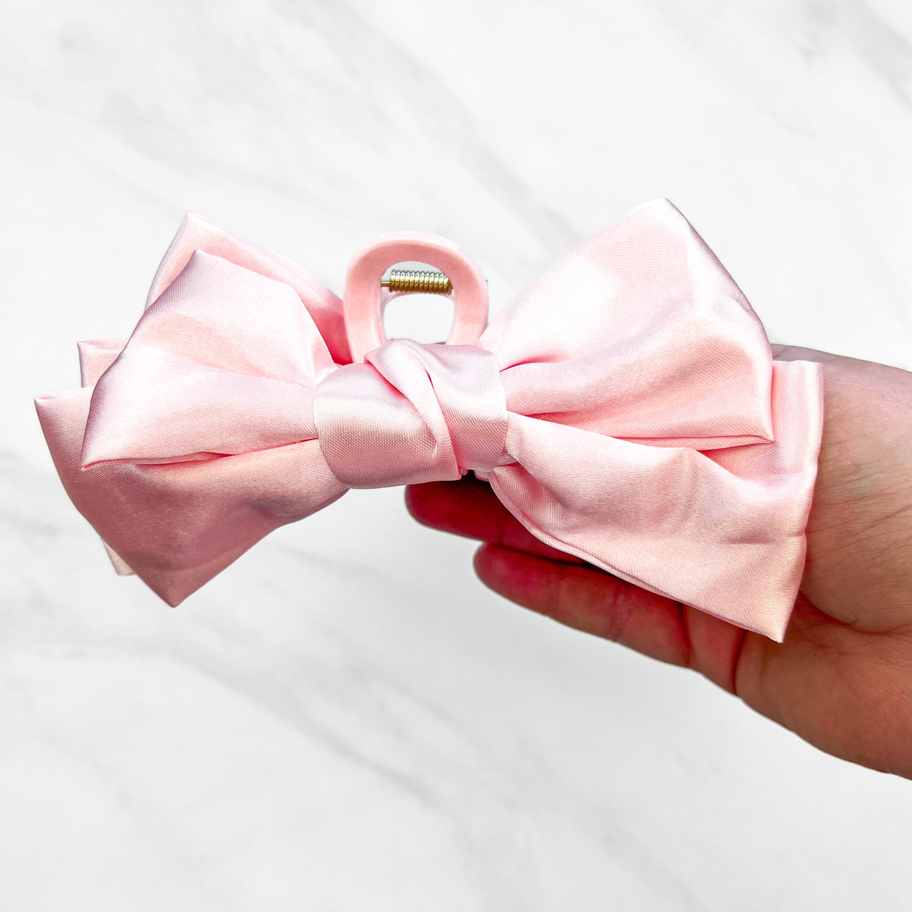 A hand holding a Classy Chic Lilac Clip, a perfect claw clip for all hair types and textures, made of silky pink satin material with a hinge closure.