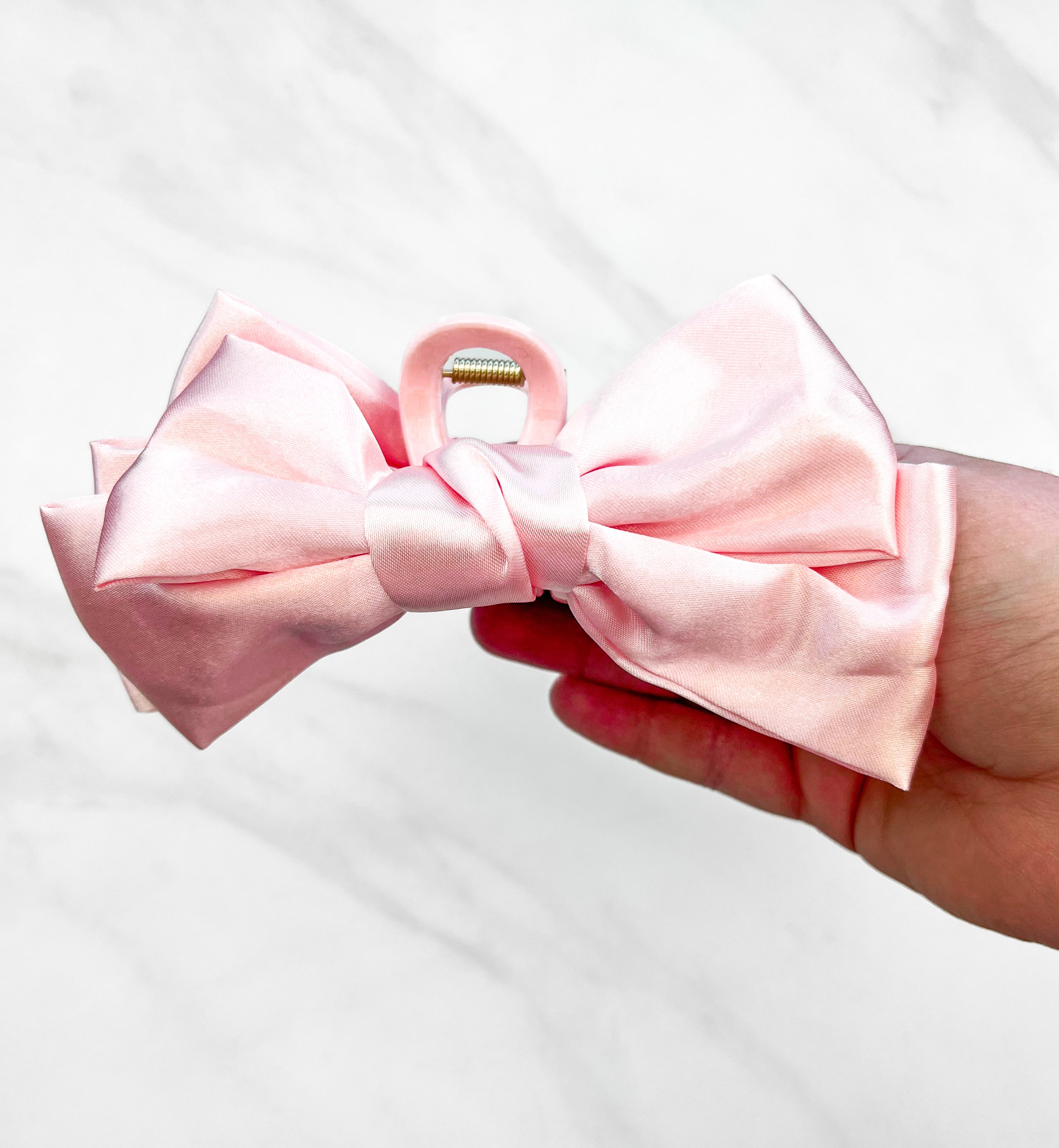 A hand holding a Classy Chic Lilac Clip, a perfect claw clip for all hair types and textures, made of silky pink satin material with a hinge closure.