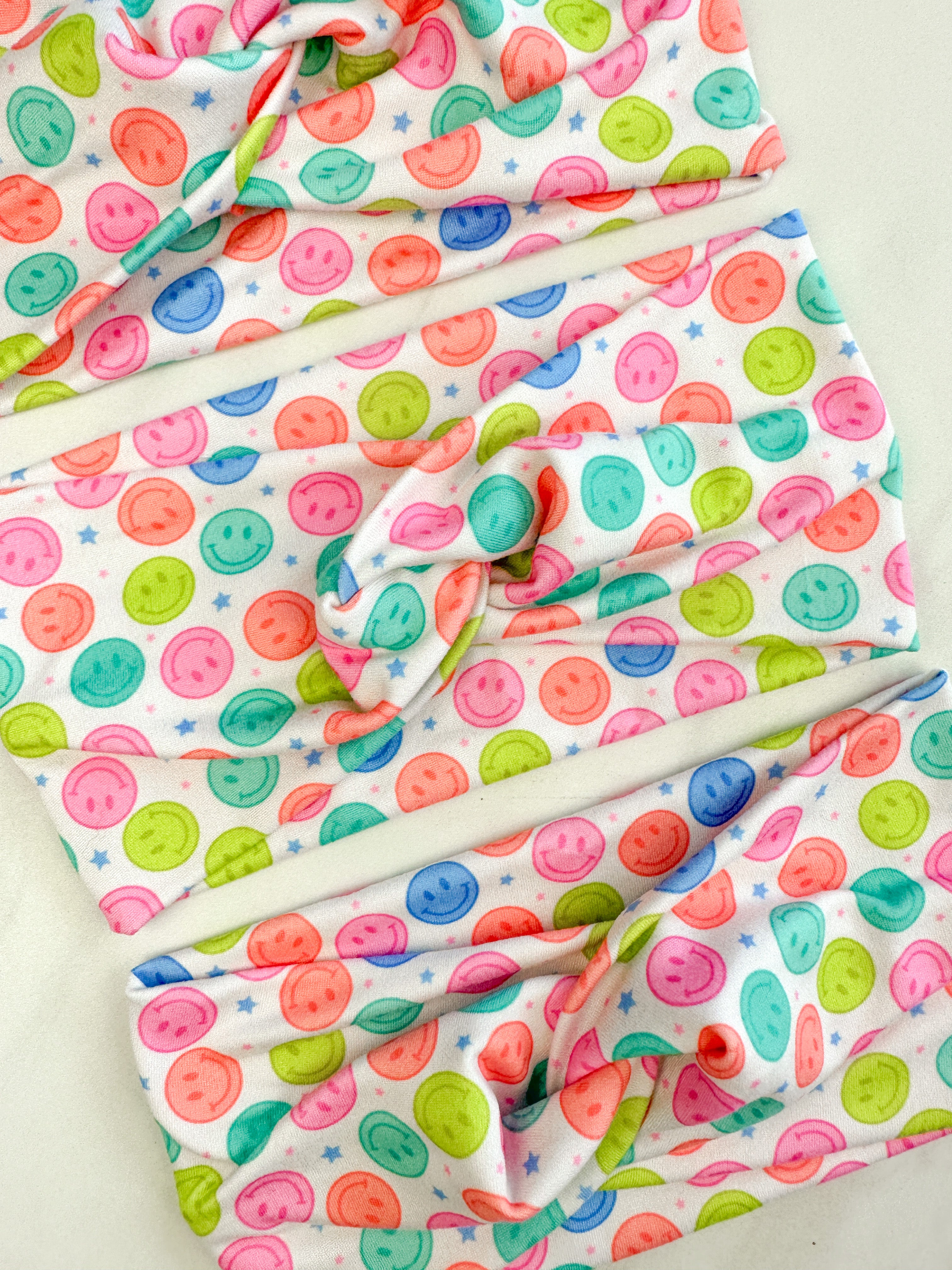Adult women's white headband with bright purple, pink, teal, coral and purple smiley faces and tiny stars