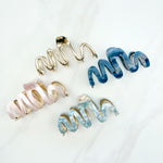 A group of zig zag-shaped marble hair clips with gold trim, including pink, blue, white and dark blue clips and one all-gold clip.