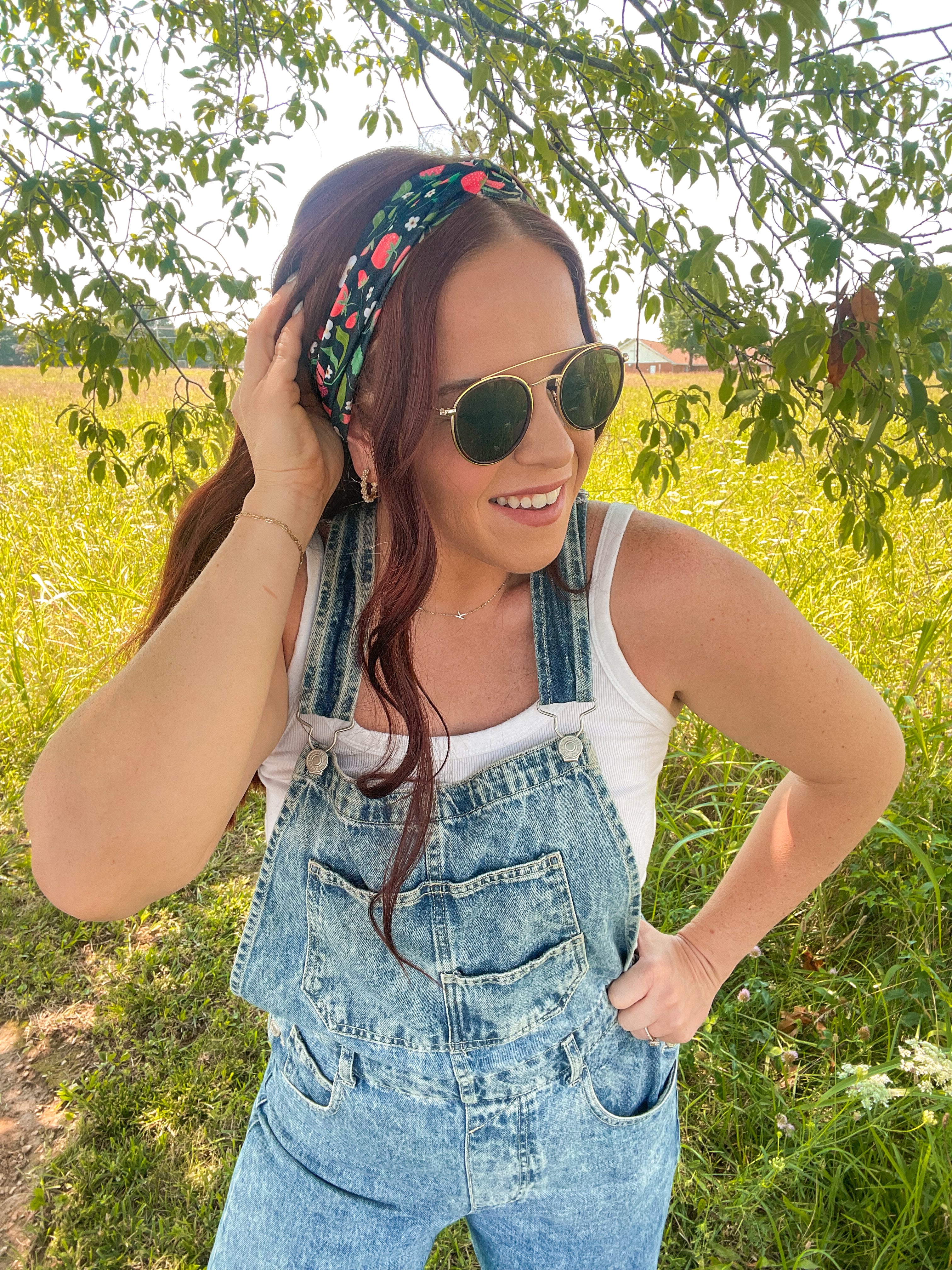 Woman in a field wearing the Feelin' Berry Good headband, sunglasses, and a white tank top with blue overalls, showcasing the headband's stylish functionality.