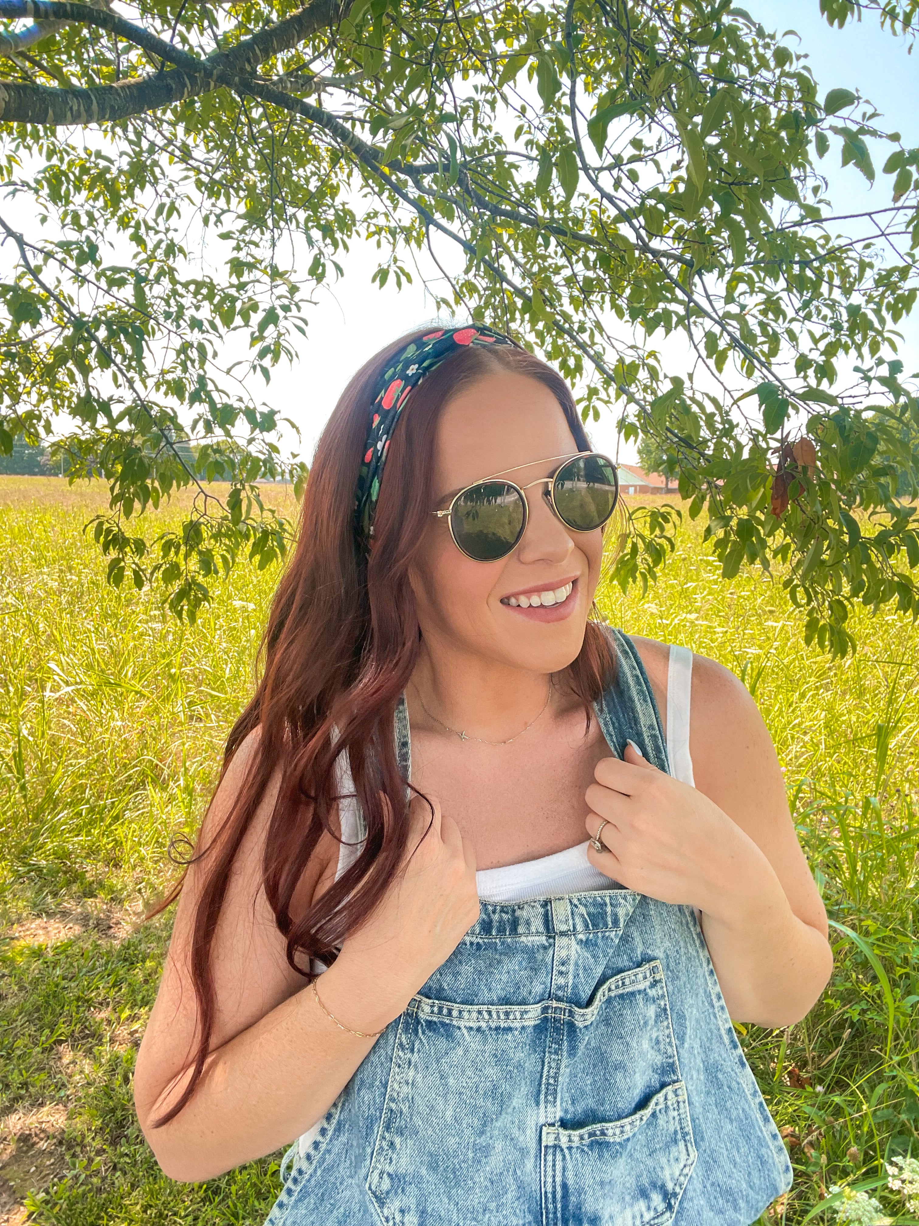 Woman in a field wearing the Feelin' Berry Good headband, sunglasses, and a white tank top with blue overalls, showcasing the headband's stylish functionality.