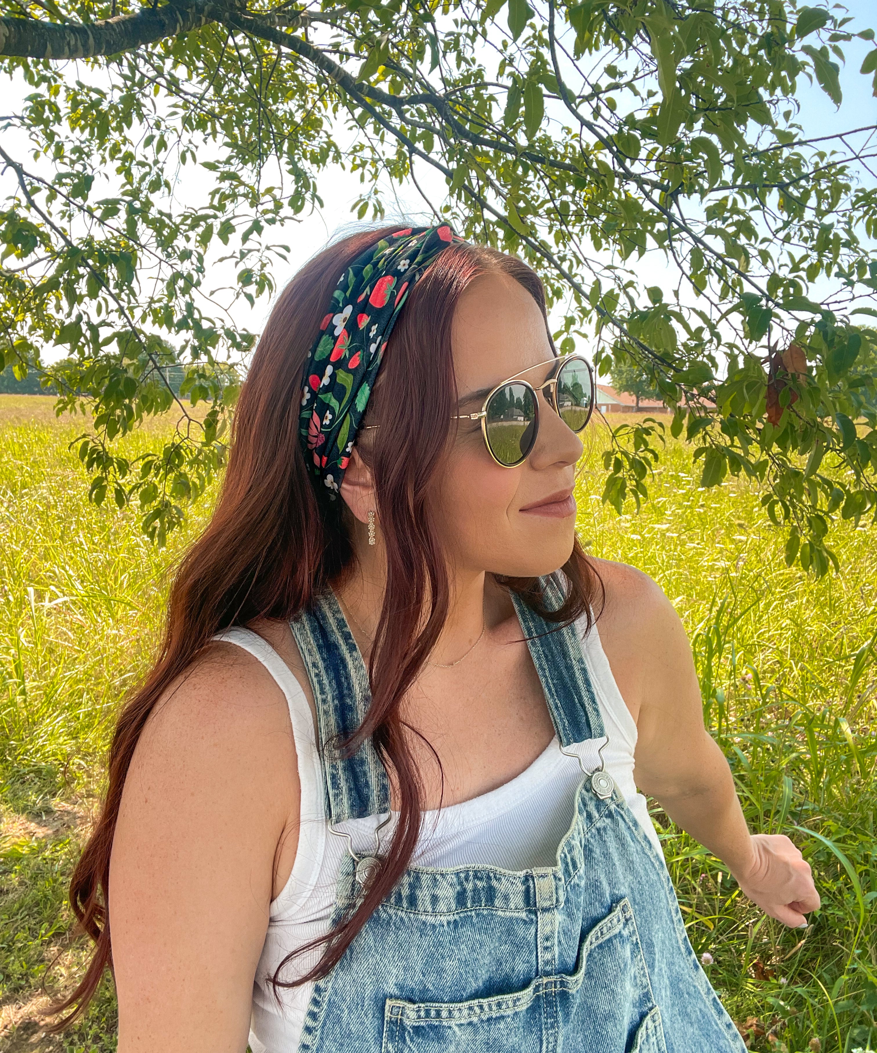 Woman in a field wearing the Feelin' Berry Good headband, sunglasses, and a white tank top with blue overalls, showcasing the headband's stylish functionality.