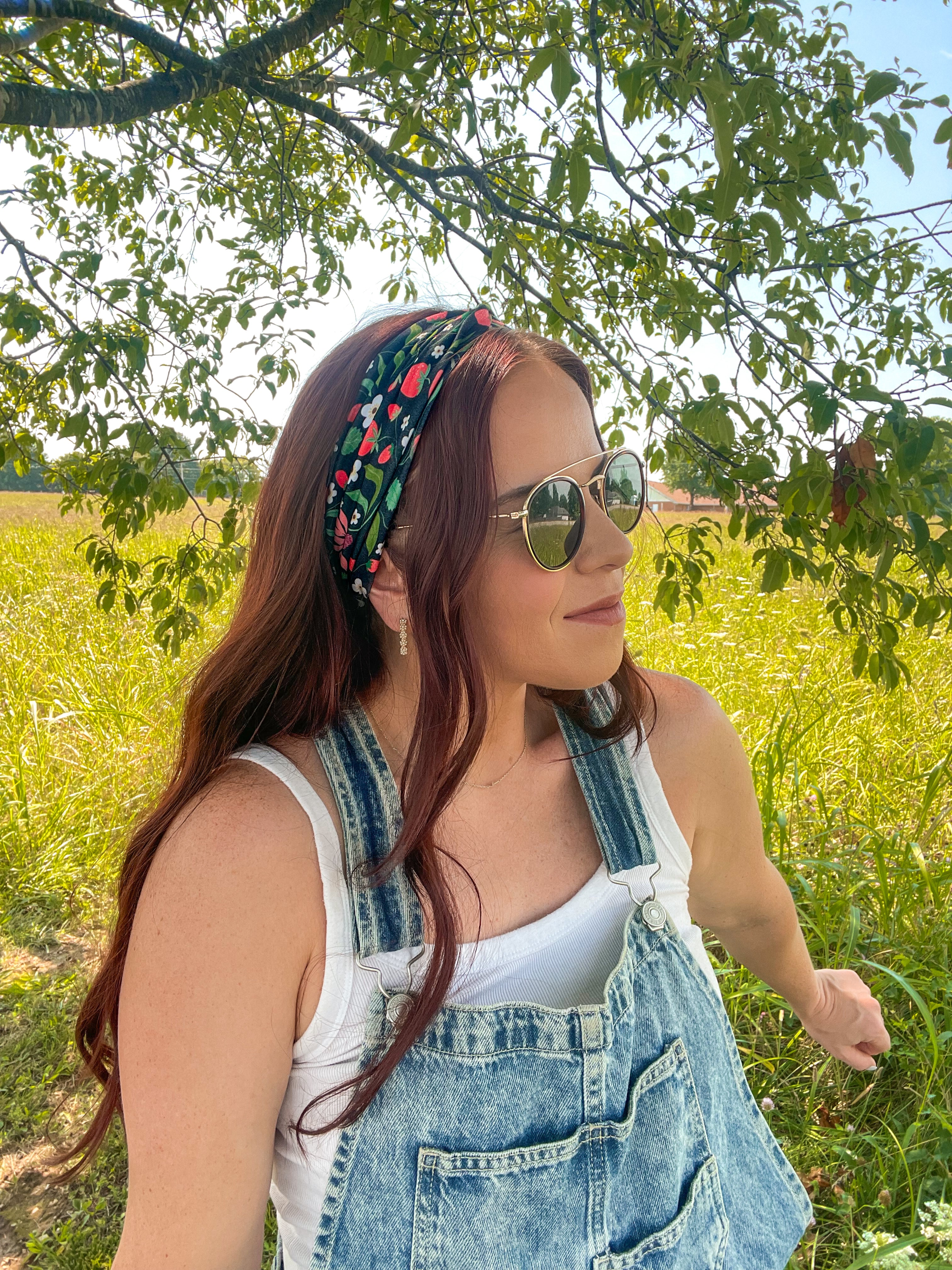 Woman in a field wearing the Feelin' Berry Good headband, sunglasses, and a white tank top with blue overalls, showcasing the headband's stylish functionality.