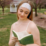 A woman confidently reads a book outdoors, wearing a Petals & Pages Lilac Market Headband. Versatile 3-in-1 twist style, sweat-absorbent, non-slip, machine washable, perfect for workouts or play. Sizes for women and children.