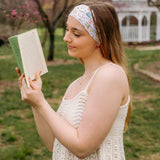 A woman confidently reads a book outdoors, wearing a Petals & Pages Lilac Market Headband. Versatile 3-in-1 twist style, sweat-absorbent, non-slip, machine washable, perfect for workouts or play. Sizes for women and children.