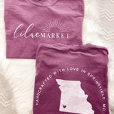 Adult berry colored shirt with Missouri state with the words "Handcrafted with Love in Springfield, MO" over the back, and "Lilac Market in script in the front with the outline of the setting sun