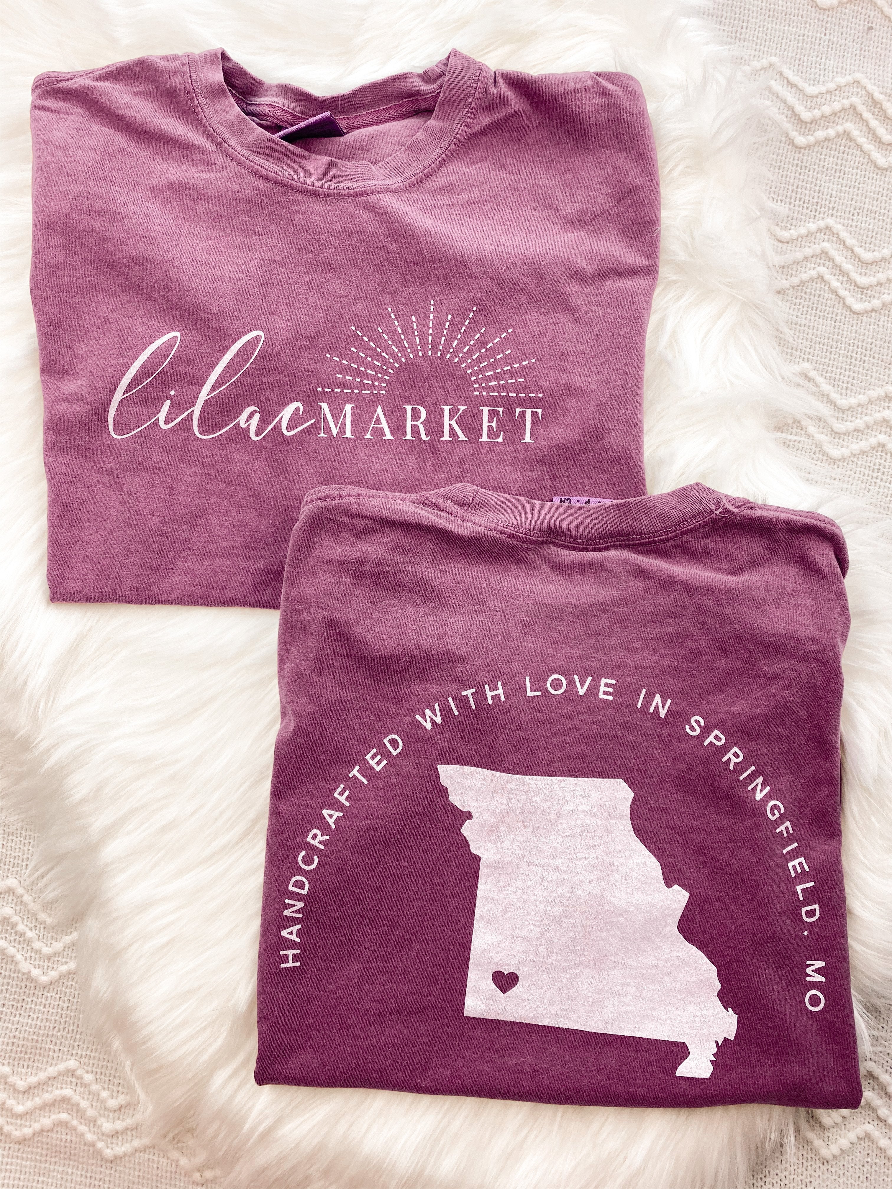 Adult berry colored shirt with Missouri state with the words "Handcrafted with Love in Springfield, MO" over the back, and "Lilac Market in script in the front with the outline of the setting sun
