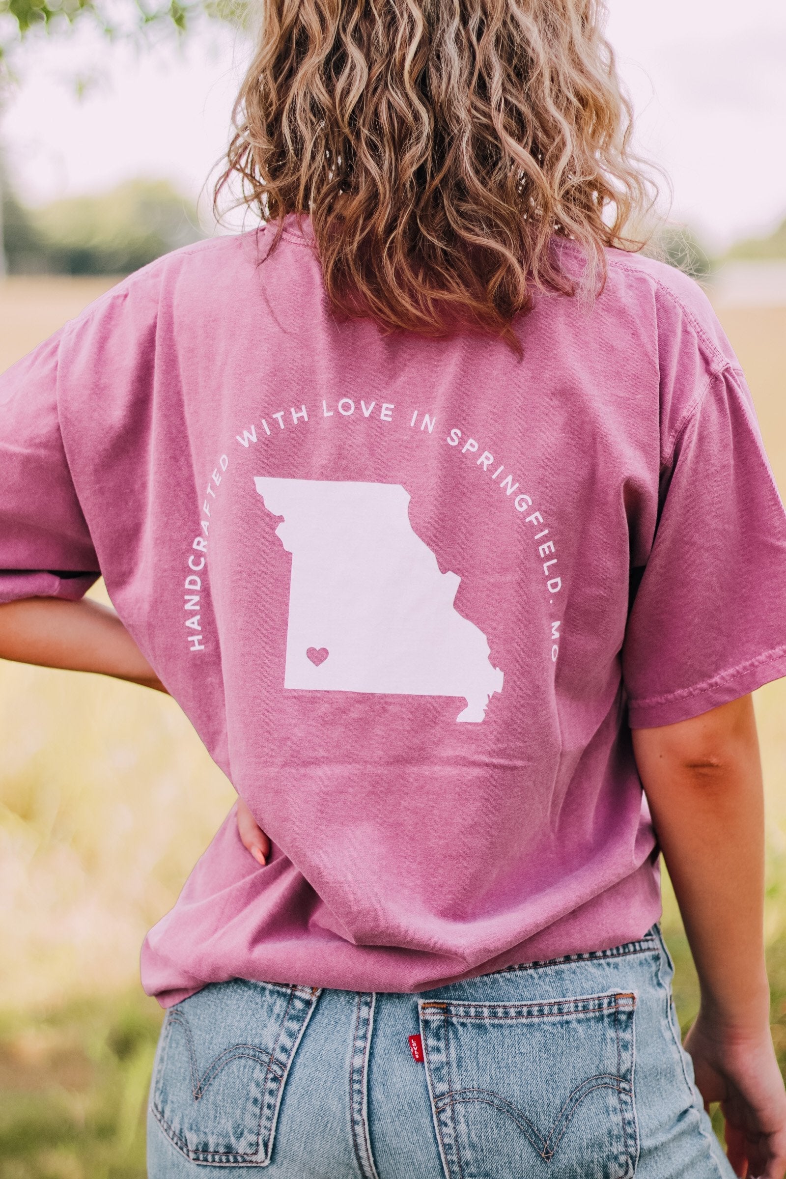 Adult berry colored shirt with Missouri state with the words "Handcrafted with Love in Springfield, MO" over the back, and "Lilac Market in script in the front with the outline of the setting sun