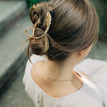 Woman wearing a gold hair clip shaped like a feather in a bun, showcasing the One of a Kind Lilac Clip from The Sarah Wallace Collection.