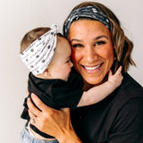 A woman smiles, holding a baby, wearing a twist knot I'm a Cool Mom - Mommy & Me headbands, adjustable for babies and toddlers. Designed in Springfield, MO.