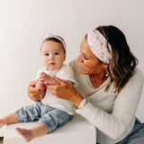 A woman smiles, holding a baby, showcasing the Sloane Floral - Mommy & Me headband set. Adjustable twist knot design for toddler and baby sizes. Designed in Springfield, MO.