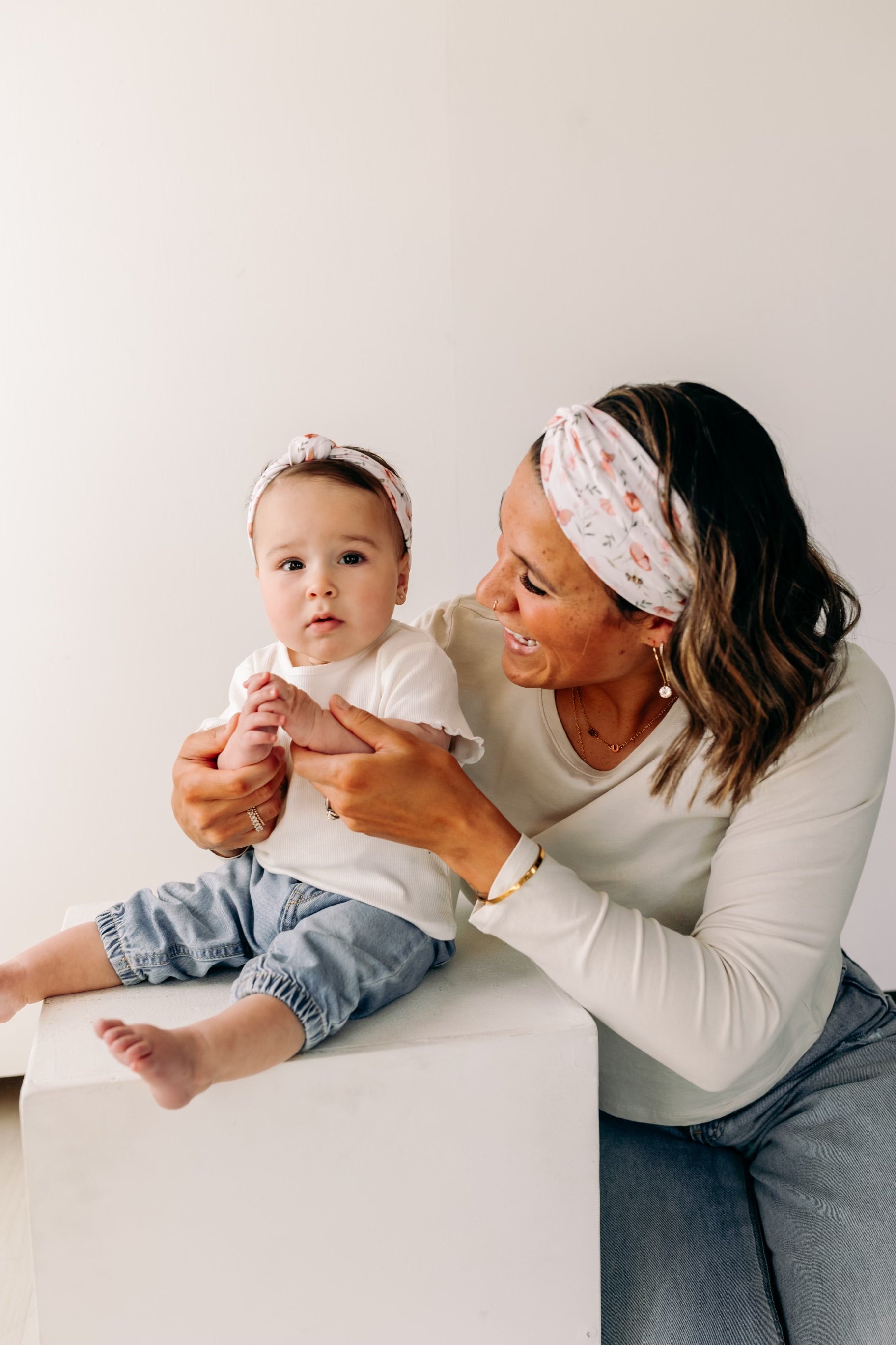 A woman smiles, holding a baby, showcasing the Sloane Floral - Mommy & Me headband set. Adjustable twist knot design for toddler and baby sizes. Designed in Springfield, MO.