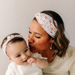 A woman smiles, holding a baby, showcasing the Sloane Floral - Mommy & Me headband set. Adjustable twist knot design for toddler and baby sizes. Designed in Springfield, MO.