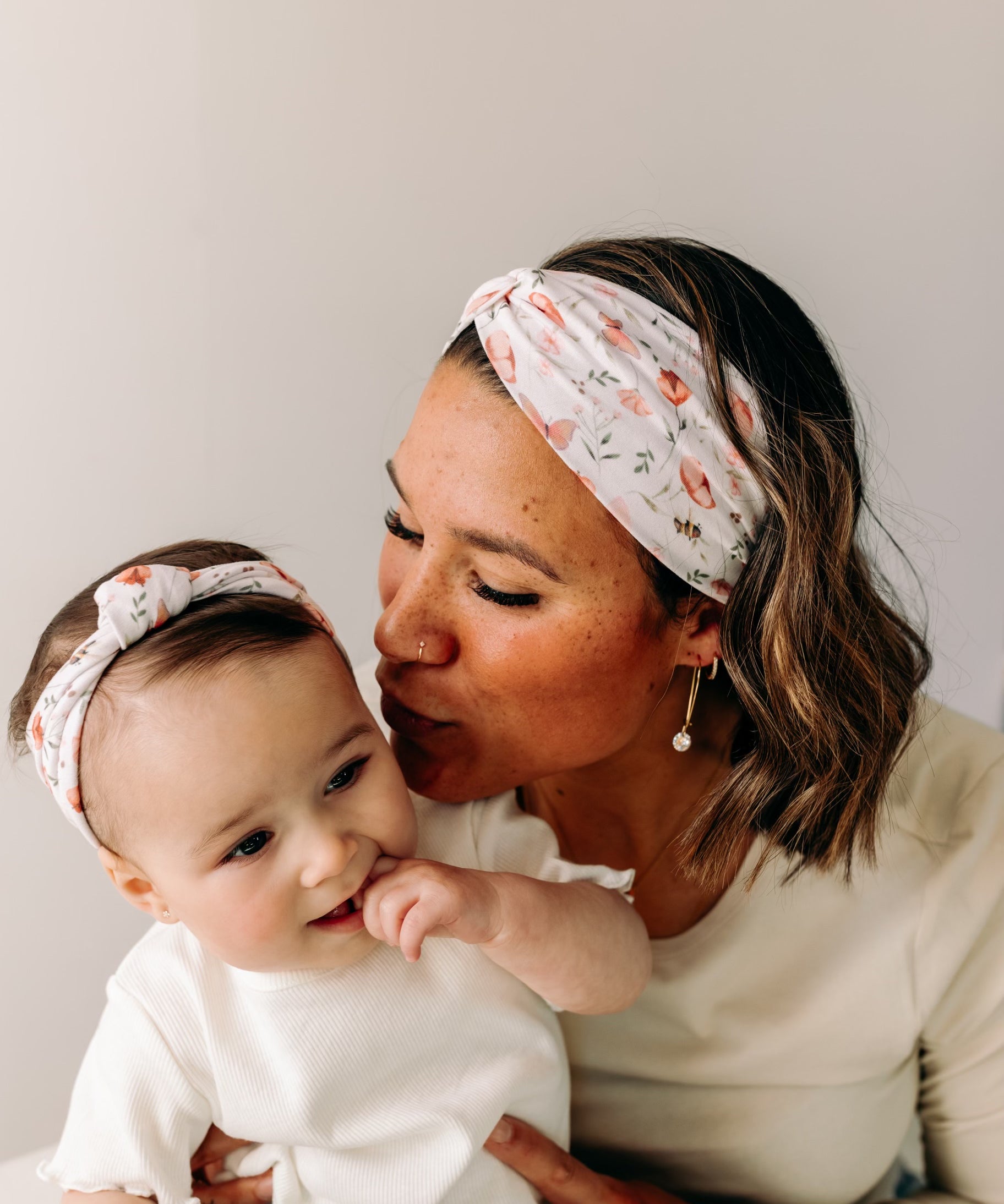A woman smiles, holding a baby, showcasing the Sloane Floral - Mommy & Me headband set. Adjustable twist knot design for toddler and baby sizes. Designed in Springfield, MO.