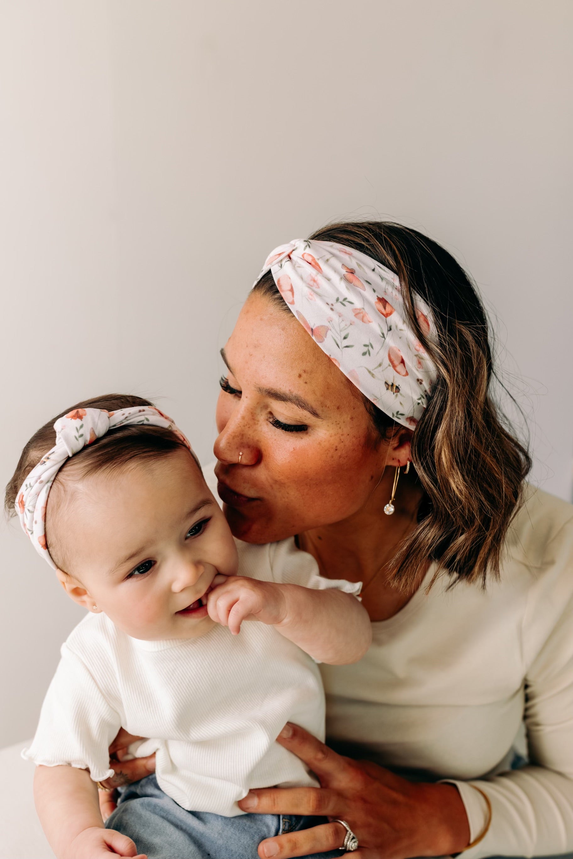 A woman smiles, holding a baby, showcasing the Sloane Floral - Mommy & Me headband set. Adjustable twist knot design for toddler and baby sizes. Designed in Springfield, MO.
