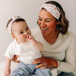 A woman smiles, holding a baby, showcasing the Sloane Floral - Mommy & Me headband set. Adjustable twist knot design for toddler and baby sizes. Designed in Springfield, MO.