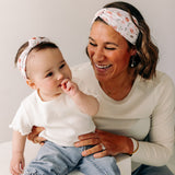 A woman smiles, holding a baby, showcasing the Sloane Floral - Mommy & Me headband set. Adjustable twist knot design for toddler and baby sizes. Designed in Springfield, MO.
