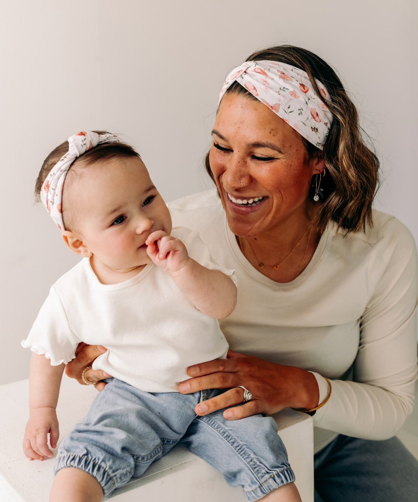 A woman smiles, holding a baby, showcasing the Sloane Floral - Mommy & Me headband set. Adjustable twist knot design for toddler and baby sizes. Designed in Springfield, MO.