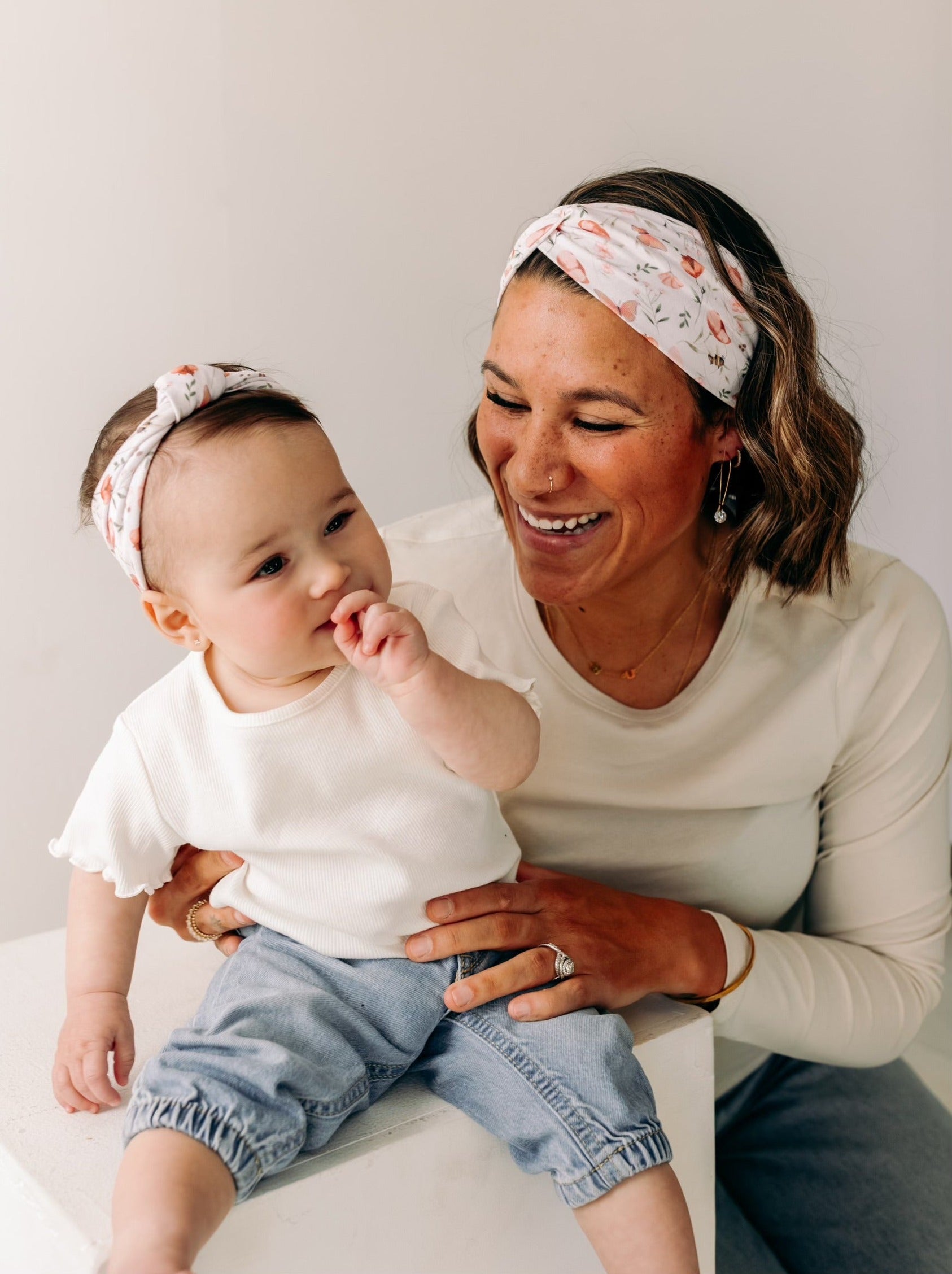A woman smiles, holding a baby, showcasing the Sloane Floral - Mommy & Me headband set. Adjustable twist knot design for toddler and baby sizes. Designed in Springfield, MO.