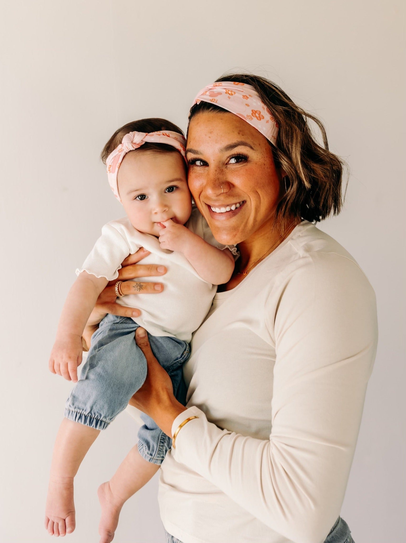 A woman smiling, holding a baby, showcasing a twist knot headband set for mommy & me. Adjustable for toddlers and babies, designed in Springfield, MO.
