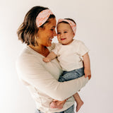 A woman smiling, holding a baby, showcasing a twist knot headband set for mommy & me. Adjustable for toddlers and babies, designed in Springfield, MO.