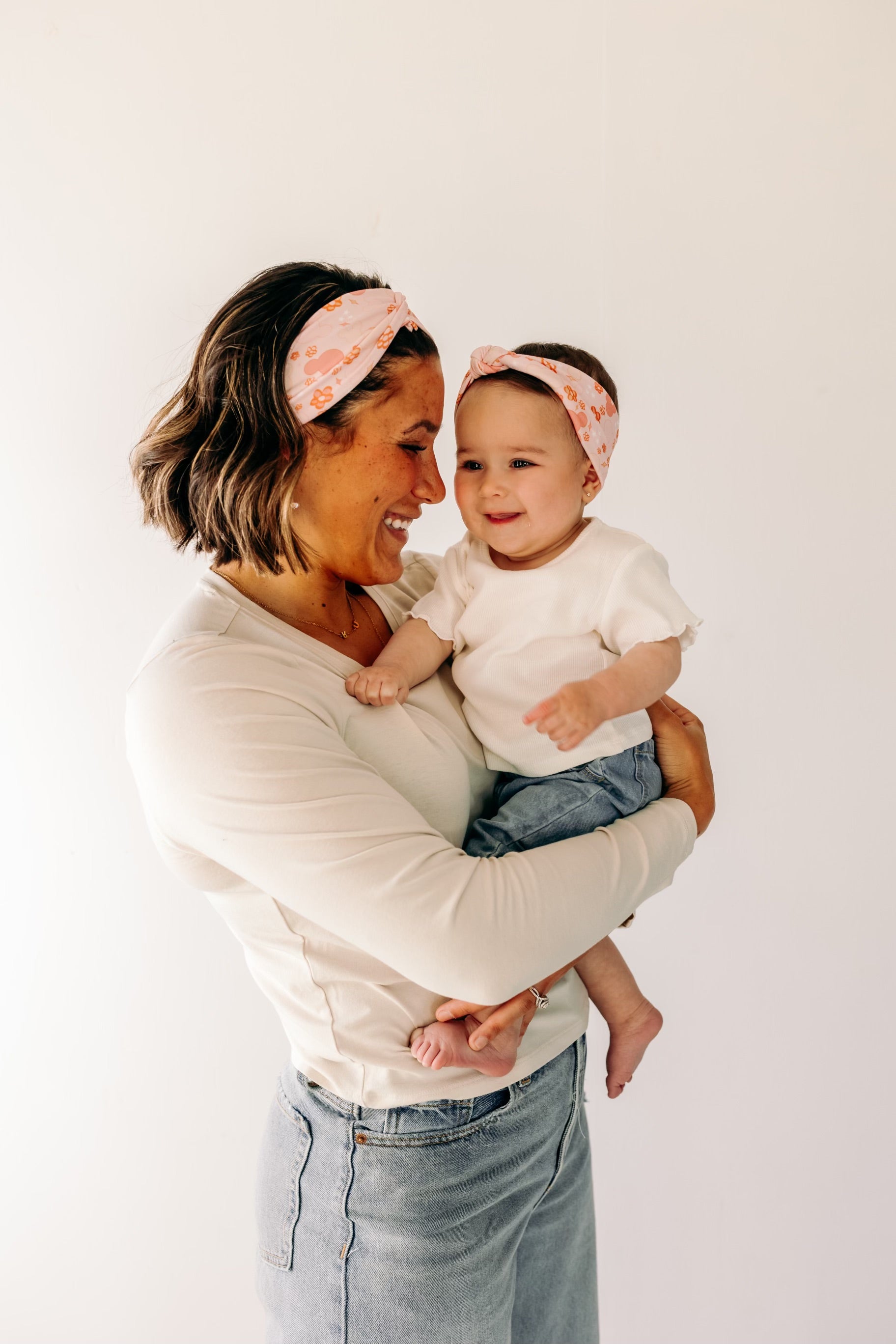 A woman smiling, holding a baby, showcasing a twist knot headband set for mommy & me. Adjustable for toddlers and babies, designed in Springfield, MO.