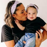 A woman smiles holding a baby, both wearing Darby Floral on Purple headbands. Adjustable bow tie style for all ages, machine washable, ultra-soft poly-knit blend. Designed in Springfield, MO.