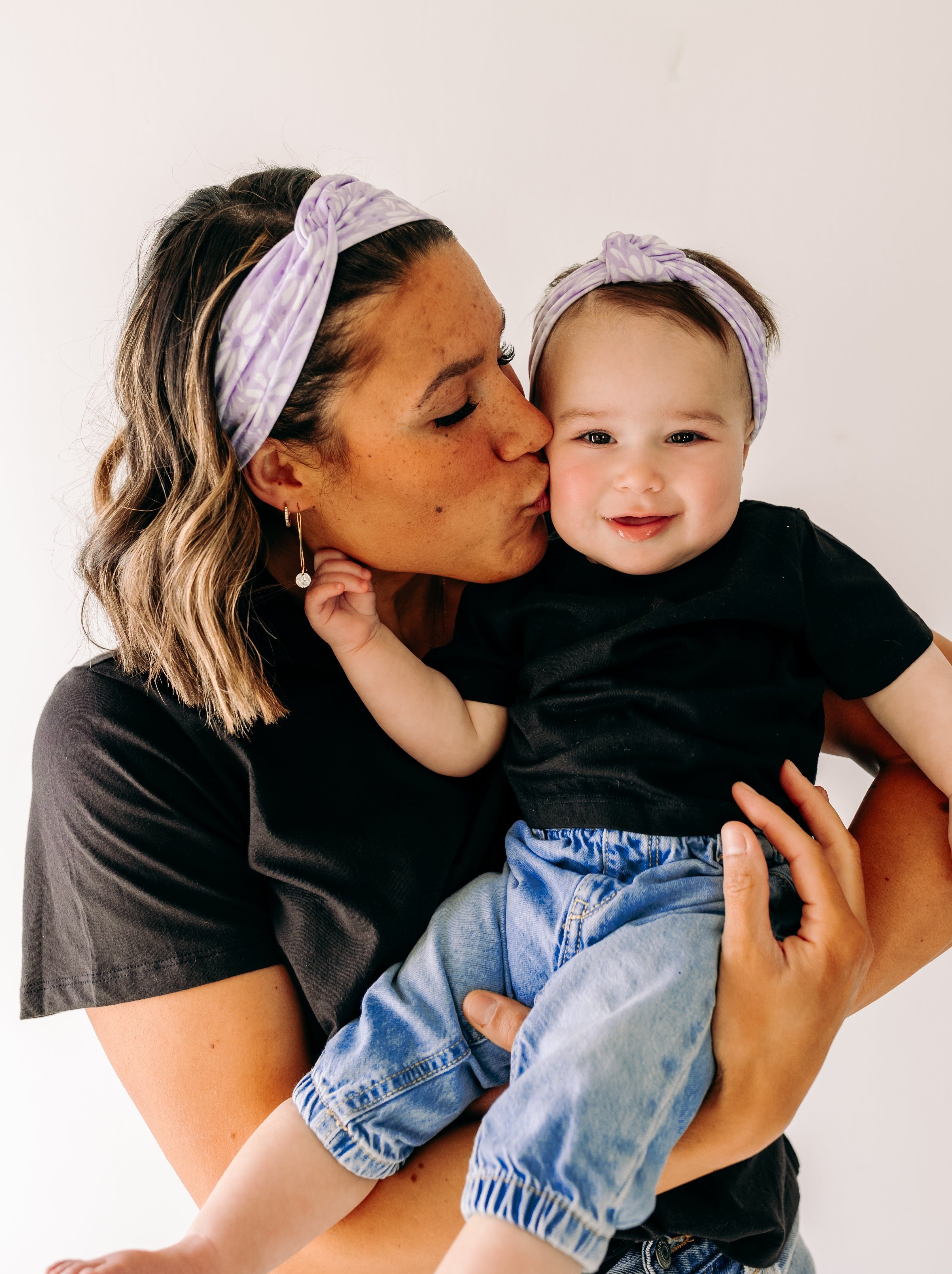 A woman smiles holding a baby, both wearing Darby Floral on Purple headbands. Adjustable bow tie style for all ages, machine washable, ultra-soft poly-knit blend. Designed in Springfield, MO.