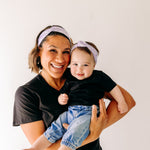 A woman smiles holding a baby, both wearing Darby Floral on Purple headbands. Adjustable bow tie style for all ages, machine washable, ultra-soft poly-knit blend. Designed in Springfield, MO.
