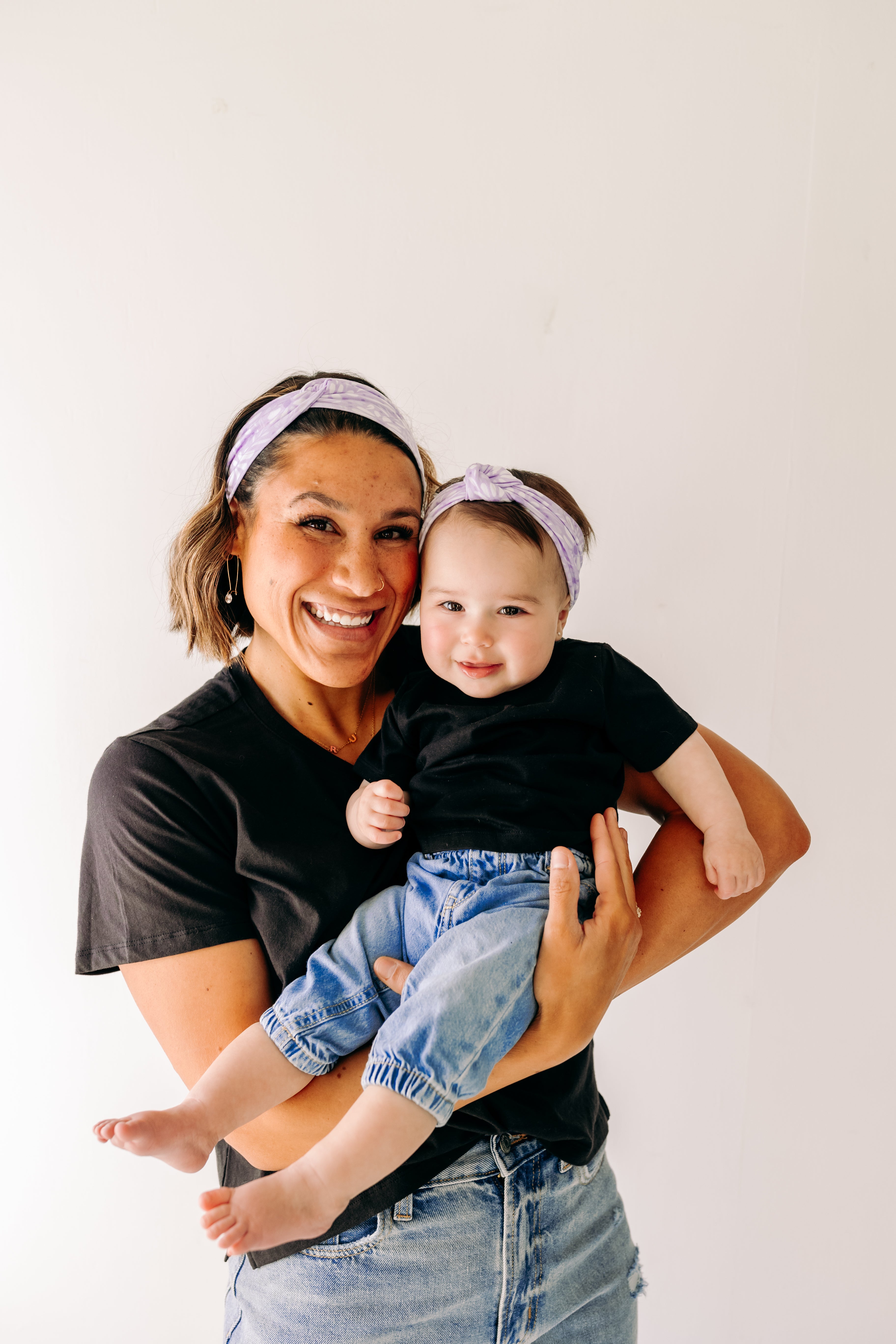 A woman smiles holding a baby, both wearing Darby Floral on Purple headbands. Adjustable bow tie style for all ages, machine washable, ultra-soft poly-knit blend. Designed in Springfield, MO.