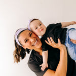 A woman smiles holding a baby, both wearing Darby Floral on Purple headbands. Adjustable bow tie style for all ages, machine washable, ultra-soft poly-knit blend. Designed in Springfield, MO.