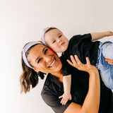 A woman smiles holding a baby, both wearing Darby Floral on Purple headbands. Adjustable bow tie style for all ages, machine washable, ultra-soft poly-knit blend. Designed in Springfield, MO.