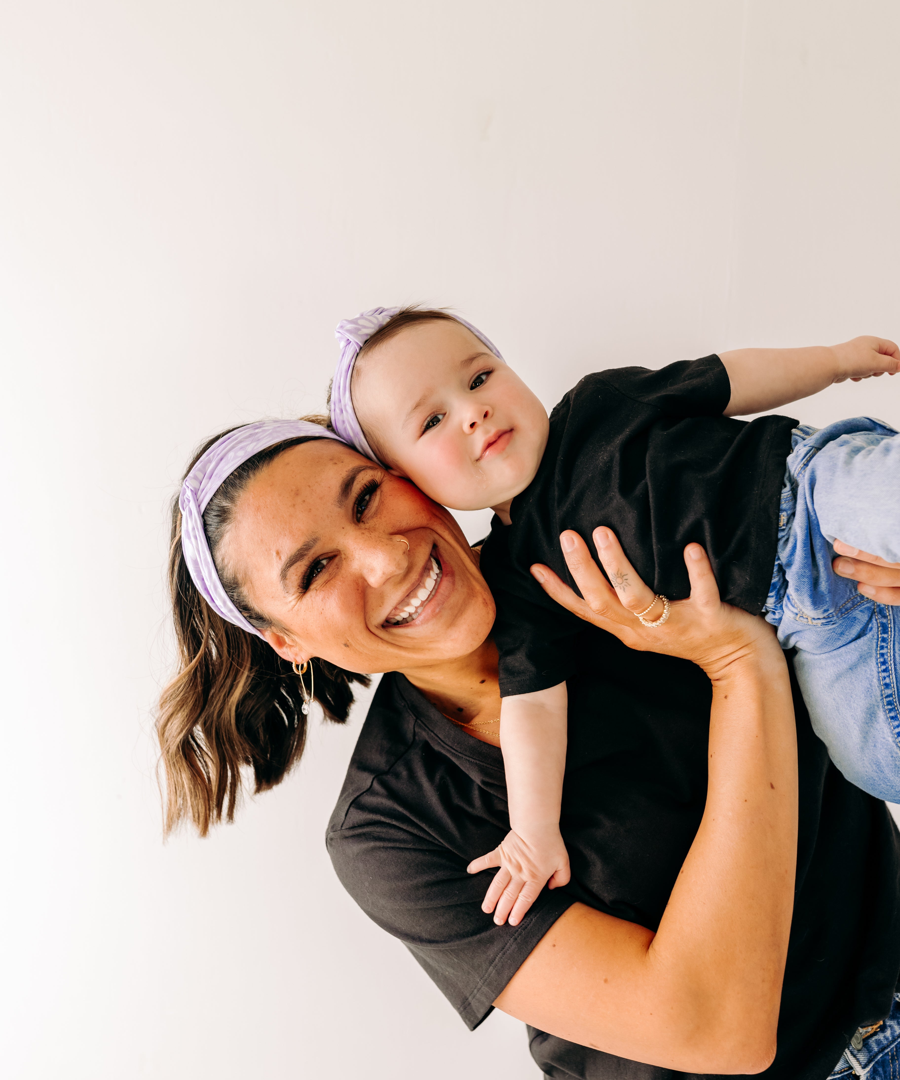 A woman smiles holding a baby, both wearing Darby Floral on Purple headbands. Adjustable bow tie style for all ages, machine washable, ultra-soft poly-knit blend. Designed in Springfield, MO.