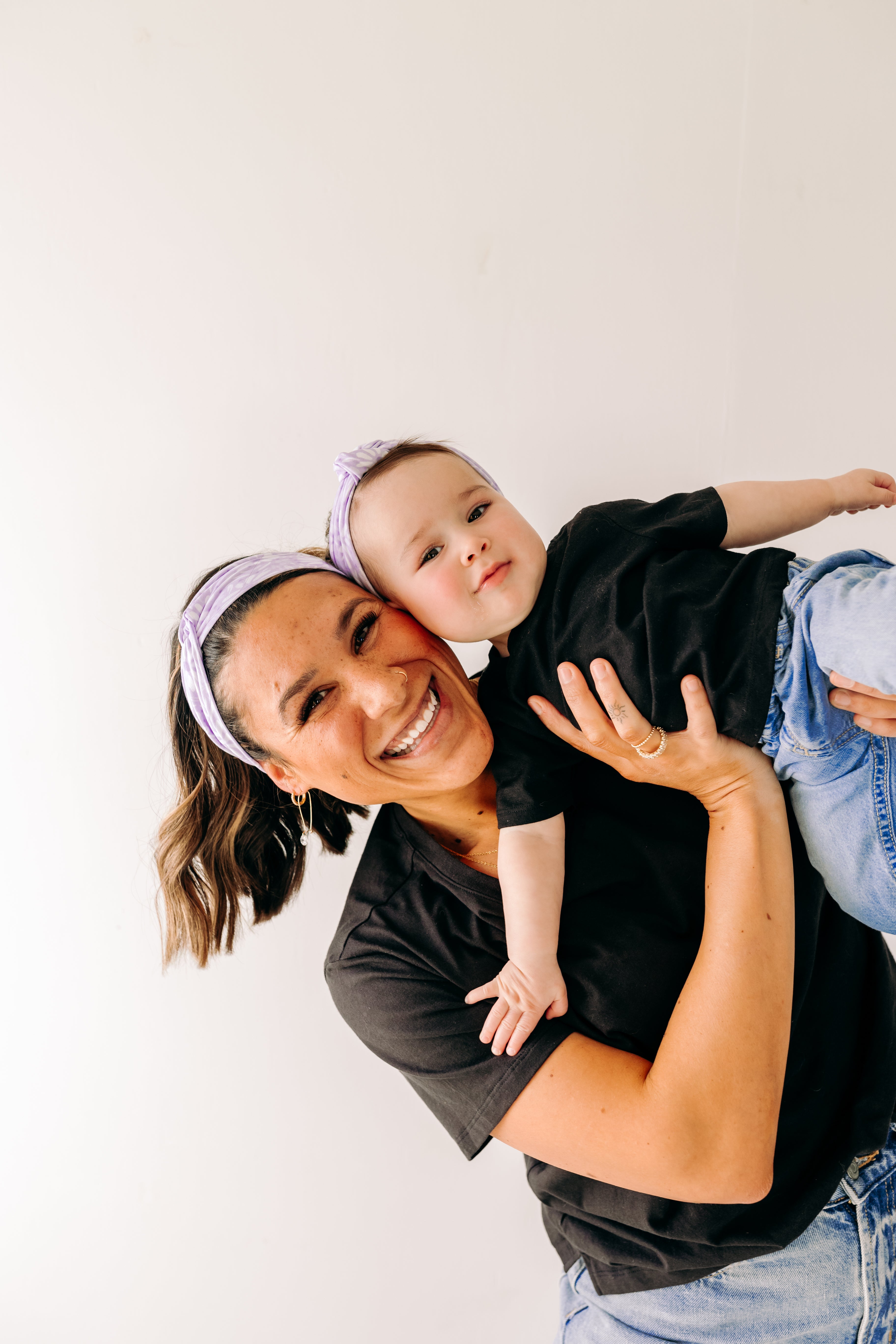 A woman smiles holding a baby, both wearing Darby Floral on Purple headbands. Adjustable bow tie style for all ages, machine washable, ultra-soft poly-knit blend. Designed in Springfield, MO.