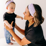 A woman smiles holding a baby, both wearing Darby Floral on Purple headbands. Adjustable bow tie style for all ages, machine washable, ultra-soft poly-knit blend. Designed in Springfield, MO.