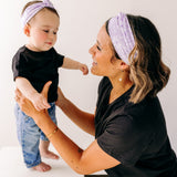 A woman smiles holding a baby, both wearing Darby Floral on Purple headbands. Adjustable bow tie style for all ages, machine washable, ultra-soft poly-knit blend. Designed in Springfield, MO.
